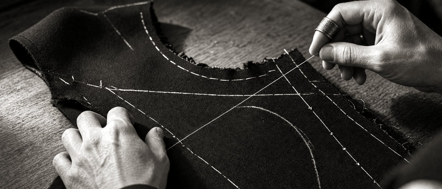 A tailor bastes the cut front of a jacket by hand, a thimble on his finger.